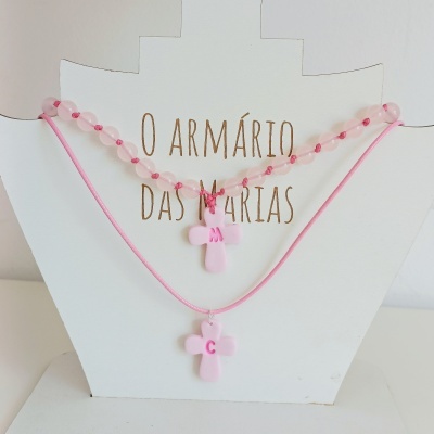 Dois colares rosa claro com pingentes de cruz e letras M e C em suporte branco com texto