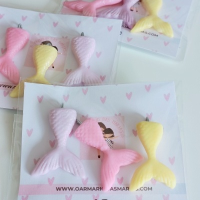 Acessórios em forma de cauda de sereia em rosa, lilás e amarelo em embalagens com padrão de corações cor de rosa.