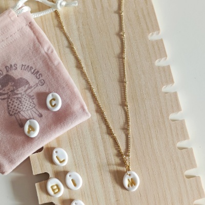 Colar dourado com pendente branco e letras douradas, letras soltas brancas com letras douradas, bolsa de pano rosa e suporte de madeira.