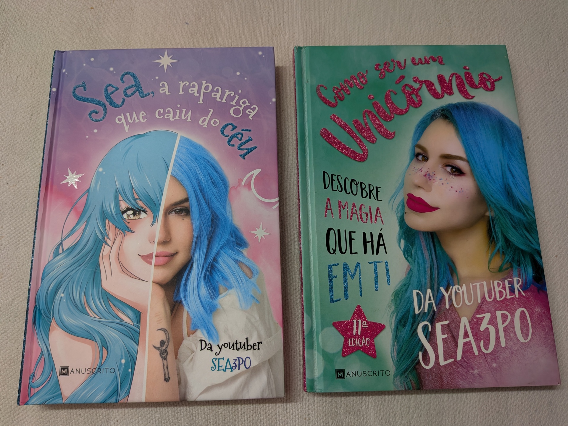 Sea3P0 - Sea, a Rapariga que Caiu do Céu + Como Ser Um Unicórnio Dois livros coloridos com capa dura, um lilás e outro verde, com imagens de mulher de cabelo azul e títulos em texto brilhante.