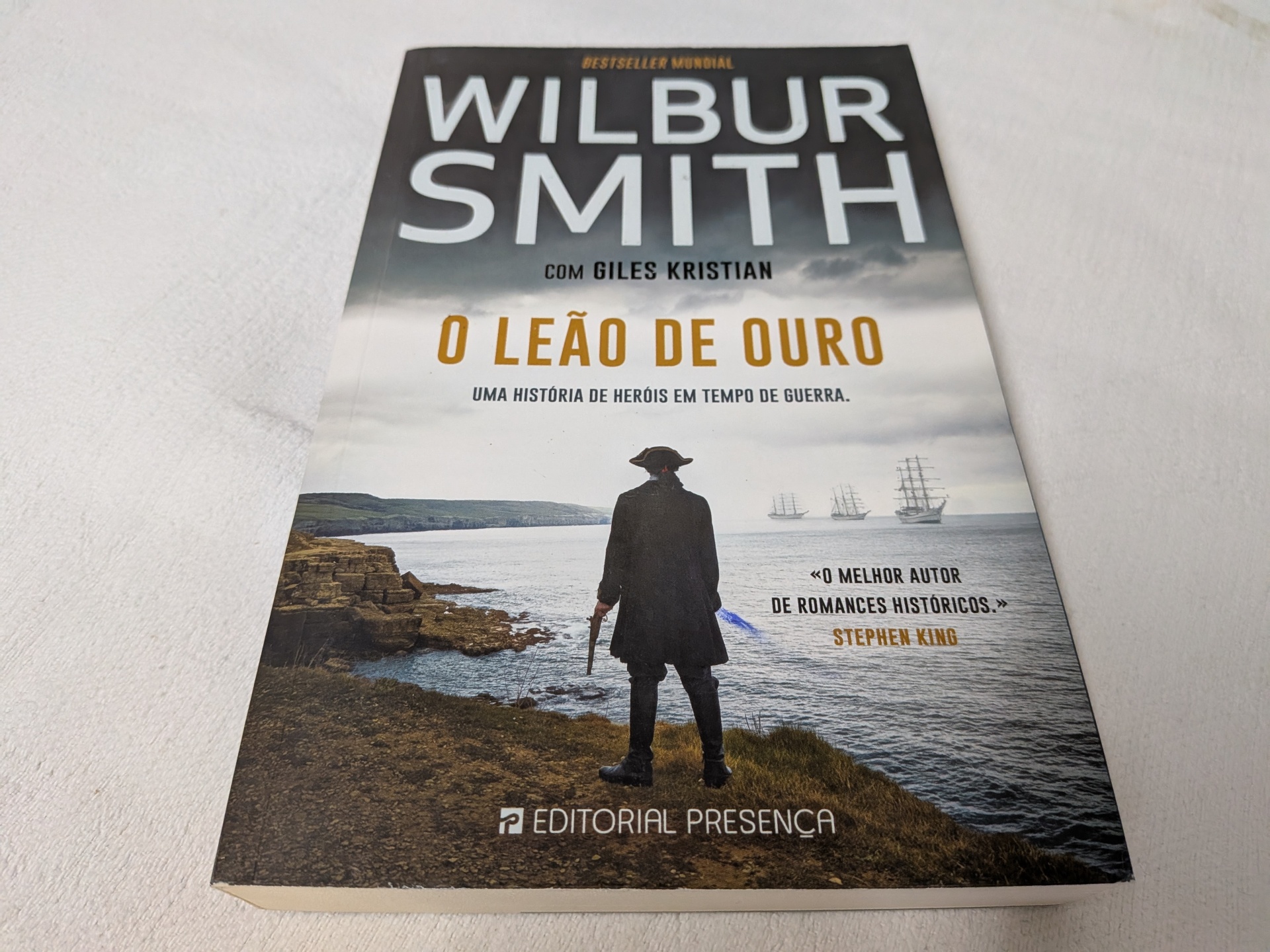 Wilbur Smith e Giles Kristian - O Leão de Ouro Capa do livro 'O Leão de Ouro' com homem na beira do mar e navios no horizonte
