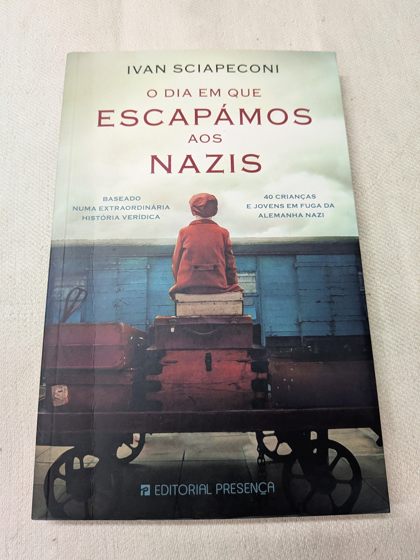 Ivan Sciapeconi - O Dia em que Escapámos aos Nazis Capa de livro com criança vestida de vermelho sentada em malas num carrinho de bagagem