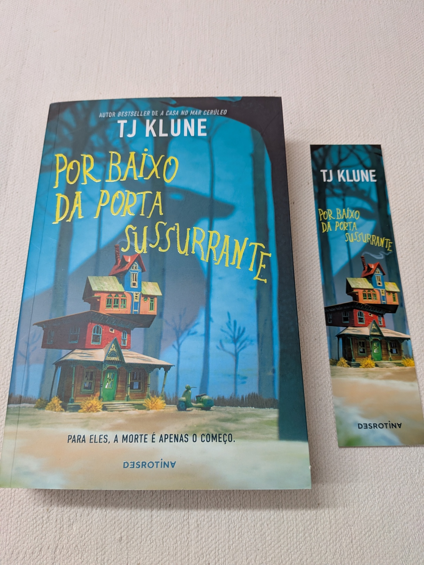 TJ Klune - Por Baixo da Porta Sussurrante Livro 'Por Baixo Da Porta Sussurrante' e marcador de livro com capa em tons azuis e amarelos com ilustração de casa e mota