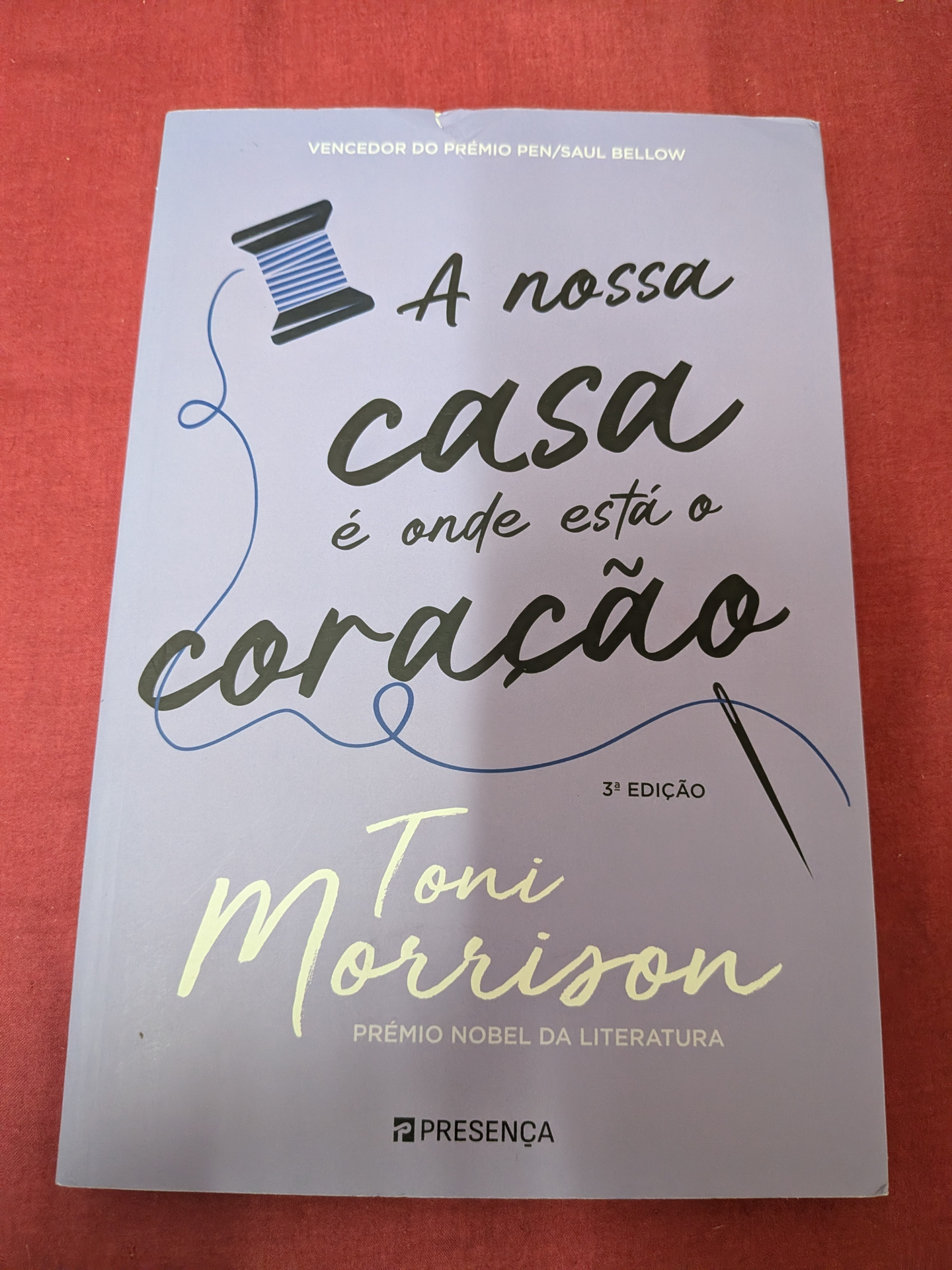 A Nossa Casa é Onde Está o Coração de Toni Morrison Capa lilás de livro A nossa casa é onde está o coração de Toni Morrison com ilustração de carretel e agulha