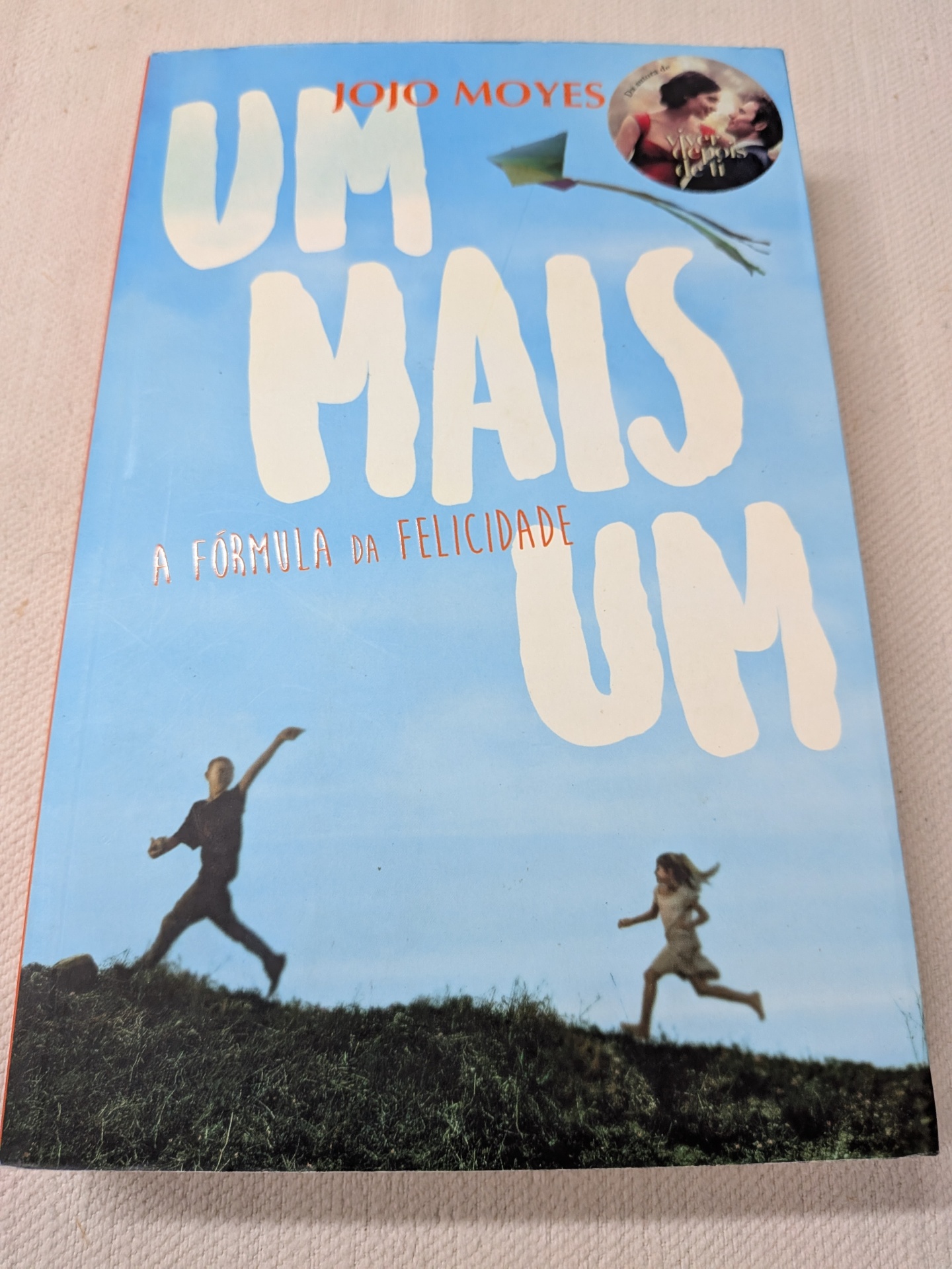 Jojo Moyes - Um Mais Um - A Fórmula da Felicidade Capa do livro Um Mais Um de Jojo Moyes com fundo azul e pessoas a correr num campo verde.