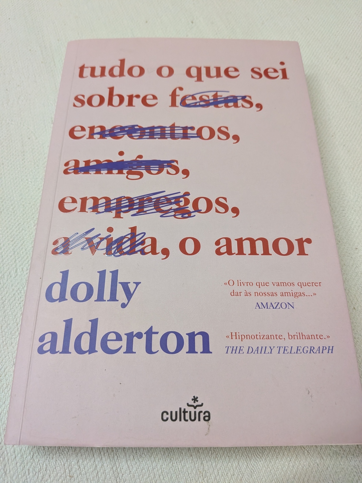 Dolly Alderton - Tudo o que Sei Sobre o Amor (Edição Limitada) Capa de livro rosa claro com título e autor em letras vermelhas e azuis, algumas palavras riscadas a caneta azul.