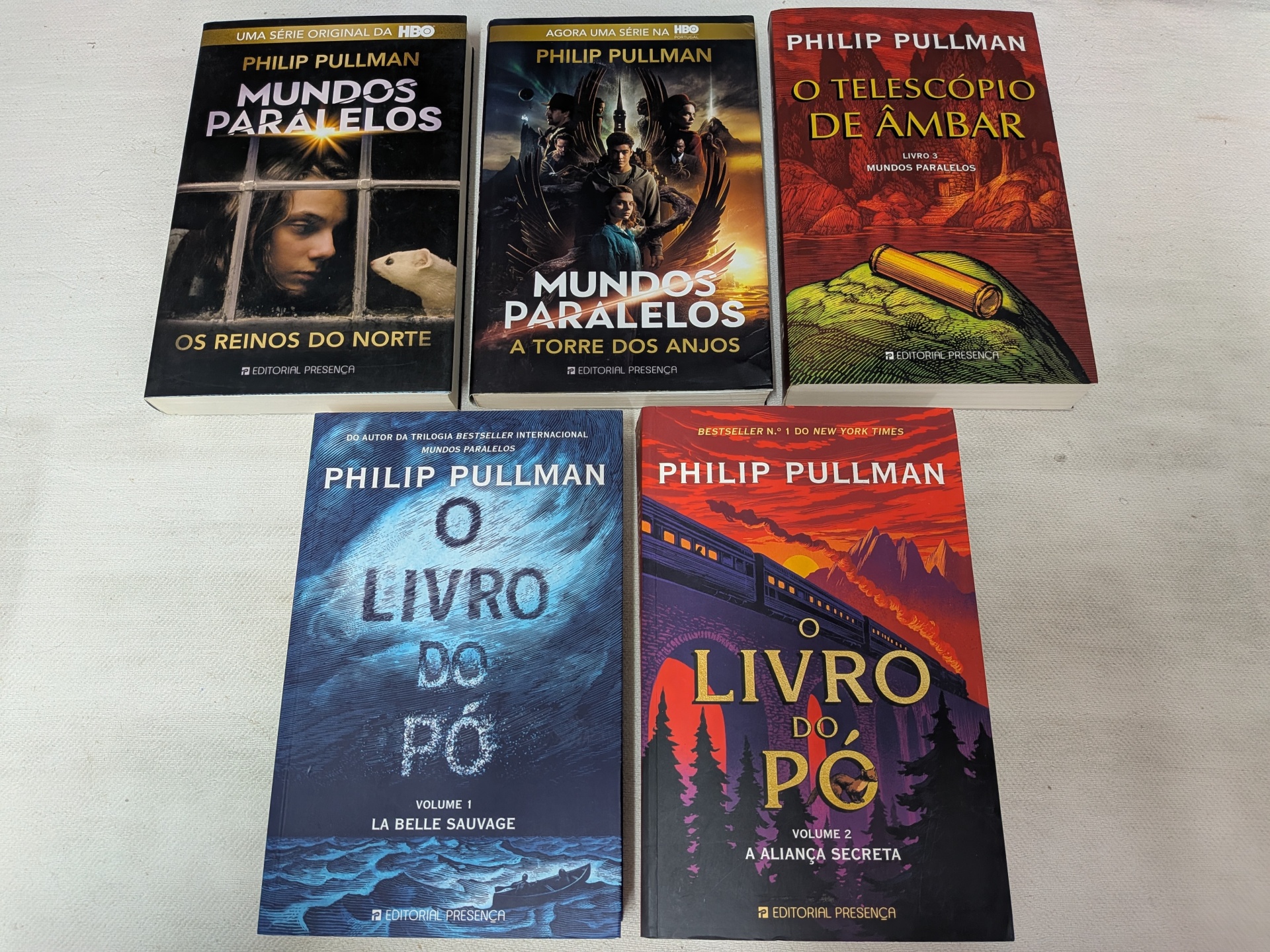 Pack Philip Pullman - Os Reinos do Norte + A Torre dos Anjos + O Telescópio de Âmbar + O Livro do Pó - Volume 1 e Volume 2 Cinco livros de Philip Pullman com títulos e capas distintas sobre uma superfície branca.
