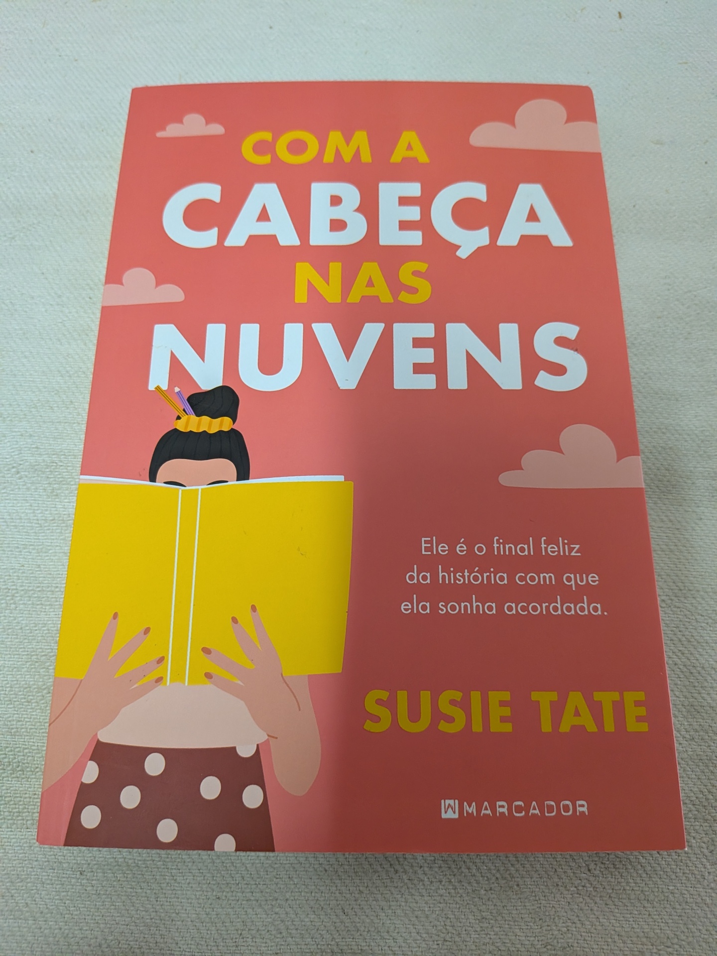 Com a Cabeça nas Nuvens de Susie Tate Capa de livro cor-de-rosa com título em letras grandes e ilustração de mulher a ler livro amarelo