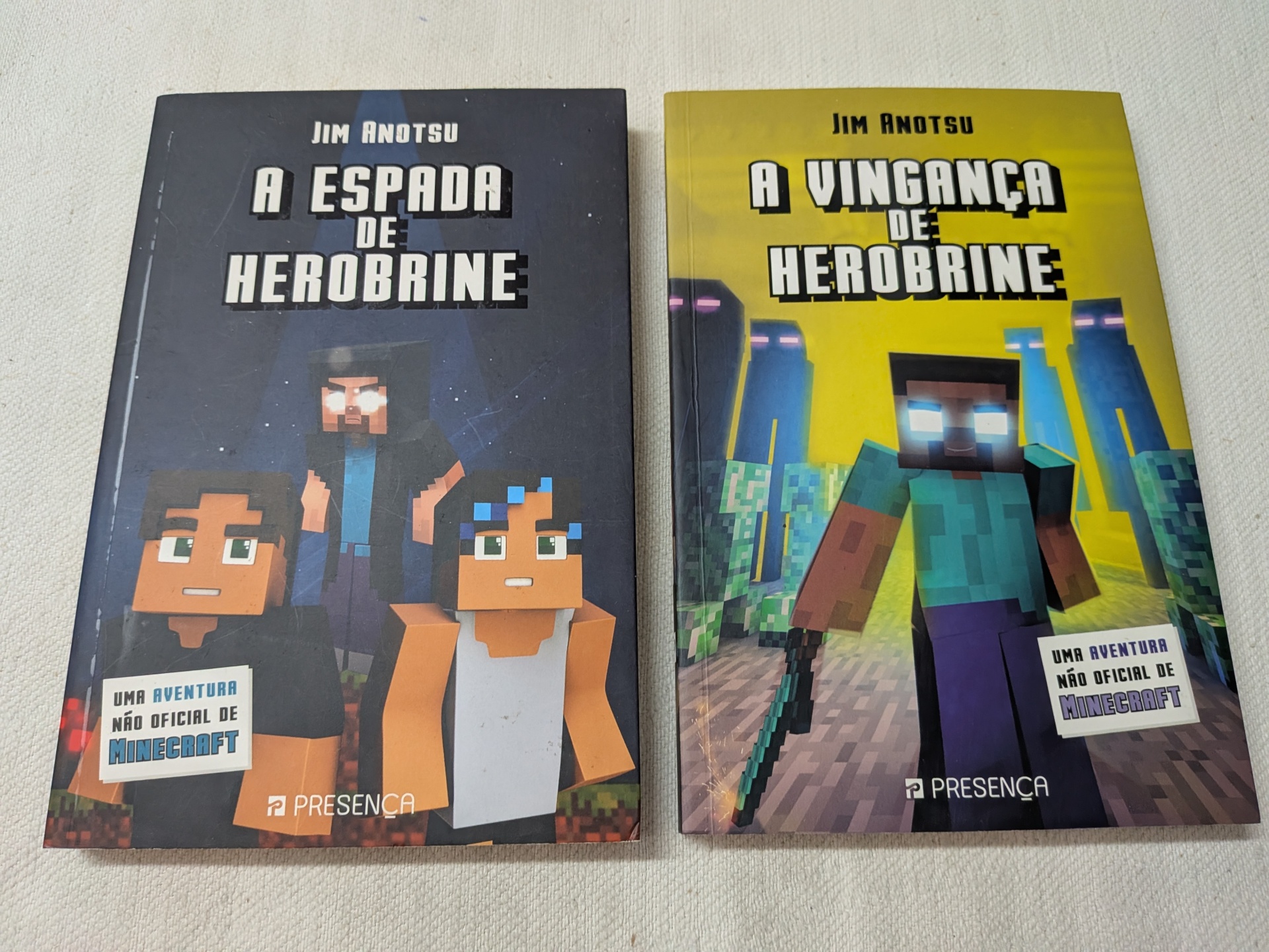 Jim Anotsu - A Espada de Herobrine + A Vingança de Herobrine Duas capas de livros estilo Minecraft com títulos em português, fundo preto e amarelo, sobre mesa clara.
