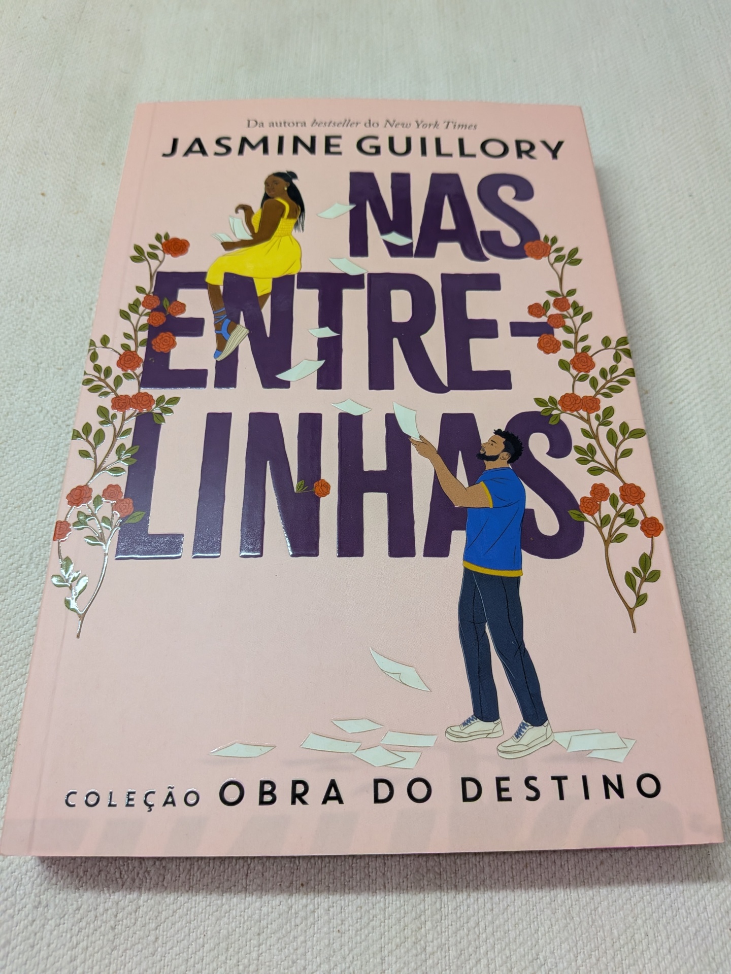 Jasmine Guillory - Nas Entrelinhas (Obra do Destino Livro 3) Capa de livro rosa claro com título NAS ENTRE-LINHAS e ilustrações de duas pessoas e flores