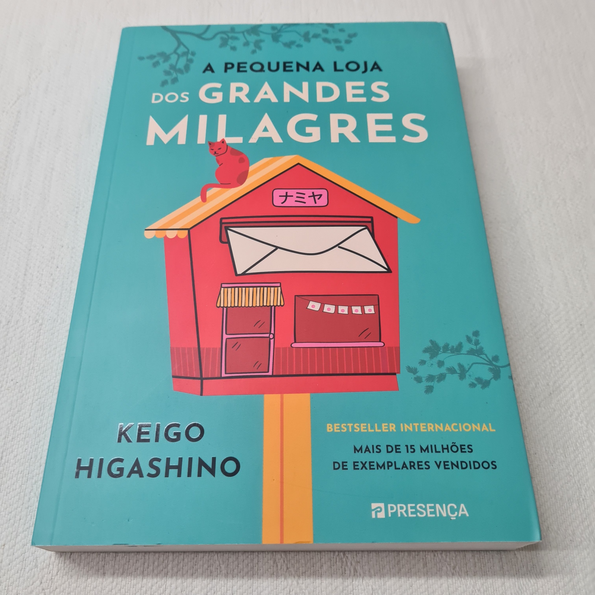 Keigo Higashino - A Pequena Loja dos Grandes Milagres Livro 'A Pequena Loja dos Grandes Milagres' com capa azul e ilustração de casinha vermelha.