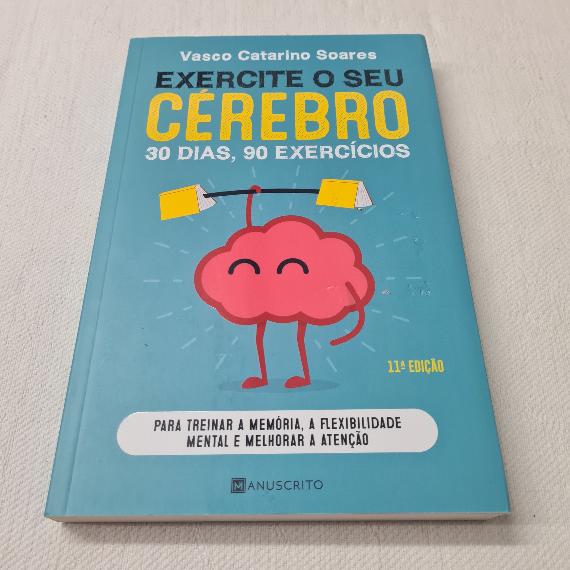 Vasco Catarino Soares - Exercite o Seu Cérebro Capa azul de livro 'Exercite o seu cérebro' com ilustração de cérebro rosa a levantar pesos amarelos