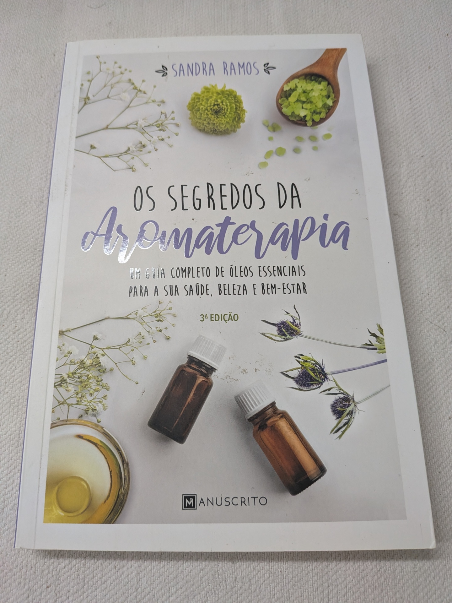 Sandra Ramos - Os Segredos da Aromaterapia Capa de livro sobre aromaterapia com frascos e flores decorativas.