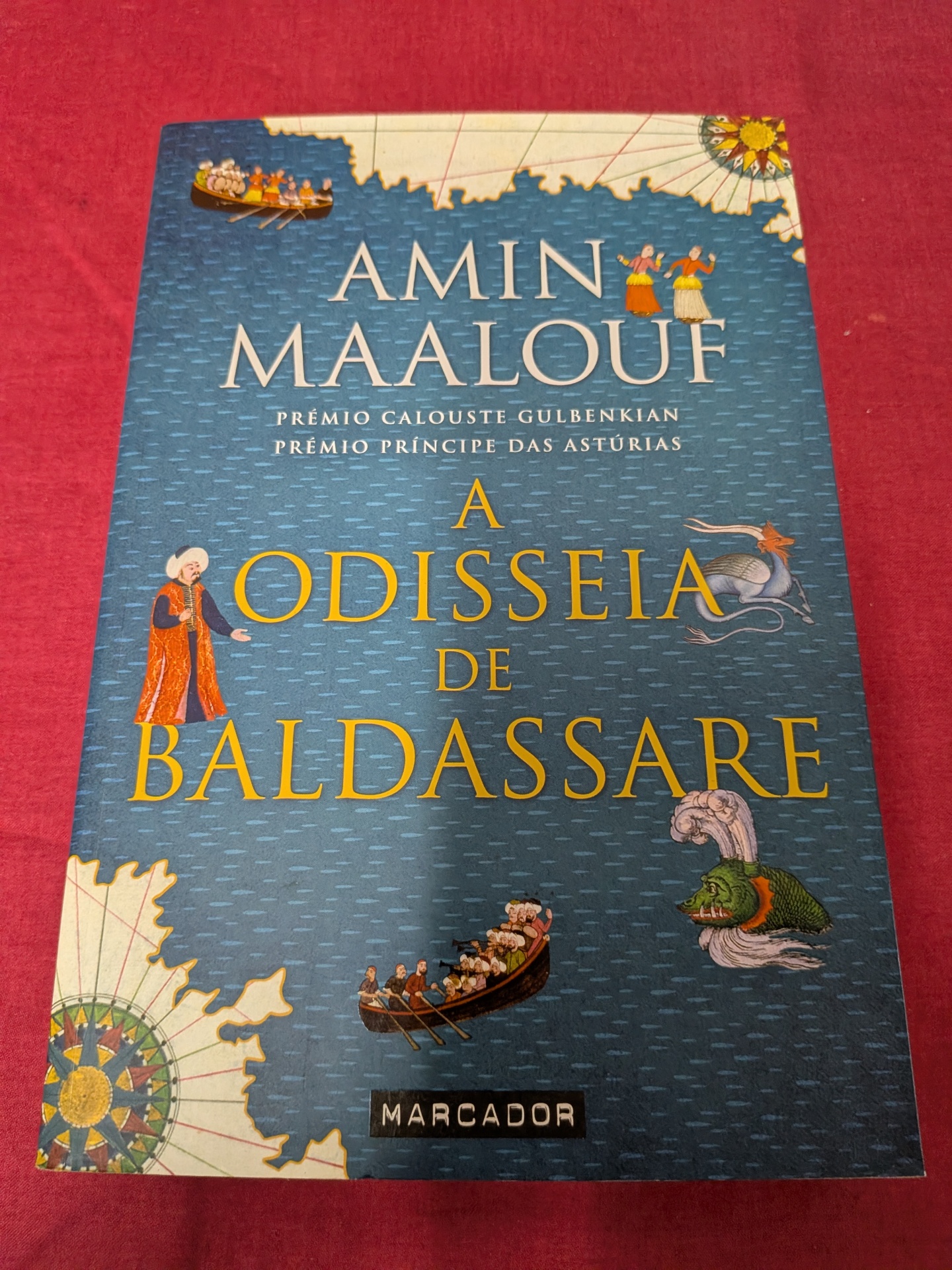 A Odisseia de Baldassare de Amin Maalouf Capa de livro azul com ilustrações e texto A ODISSEIA DE BALDASSARE.
