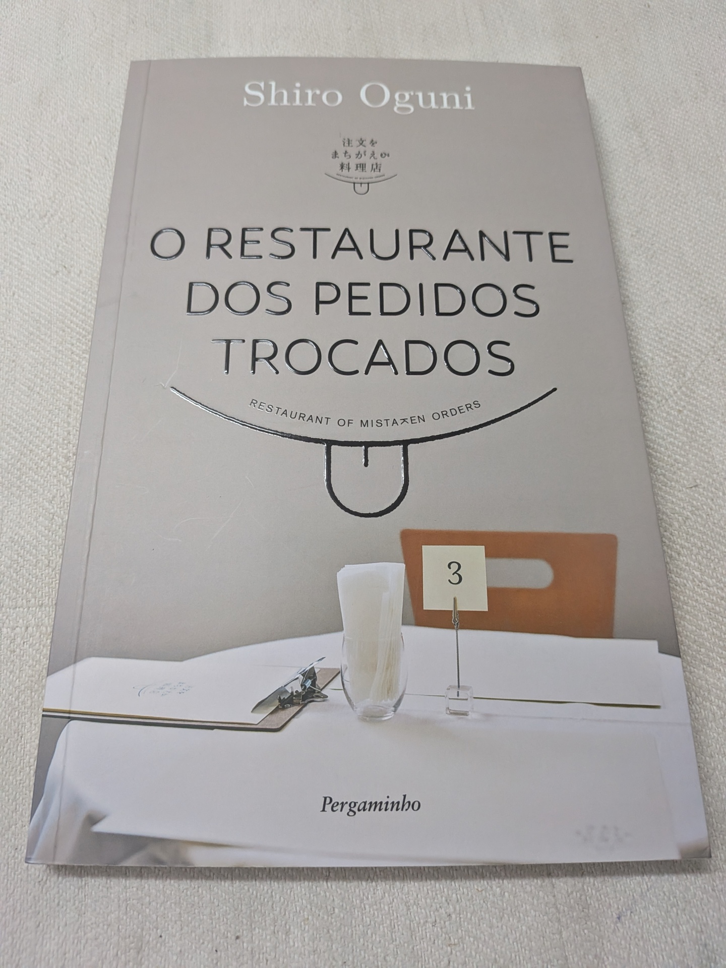 Shiro Oguni - O Restaurante dos Pedidos Trocados Capa de livro com título O RESTAURANTE DOS PEDIDOS TROCADOS e uma mesa de restaurante simples com itens sobre a toalha branca.