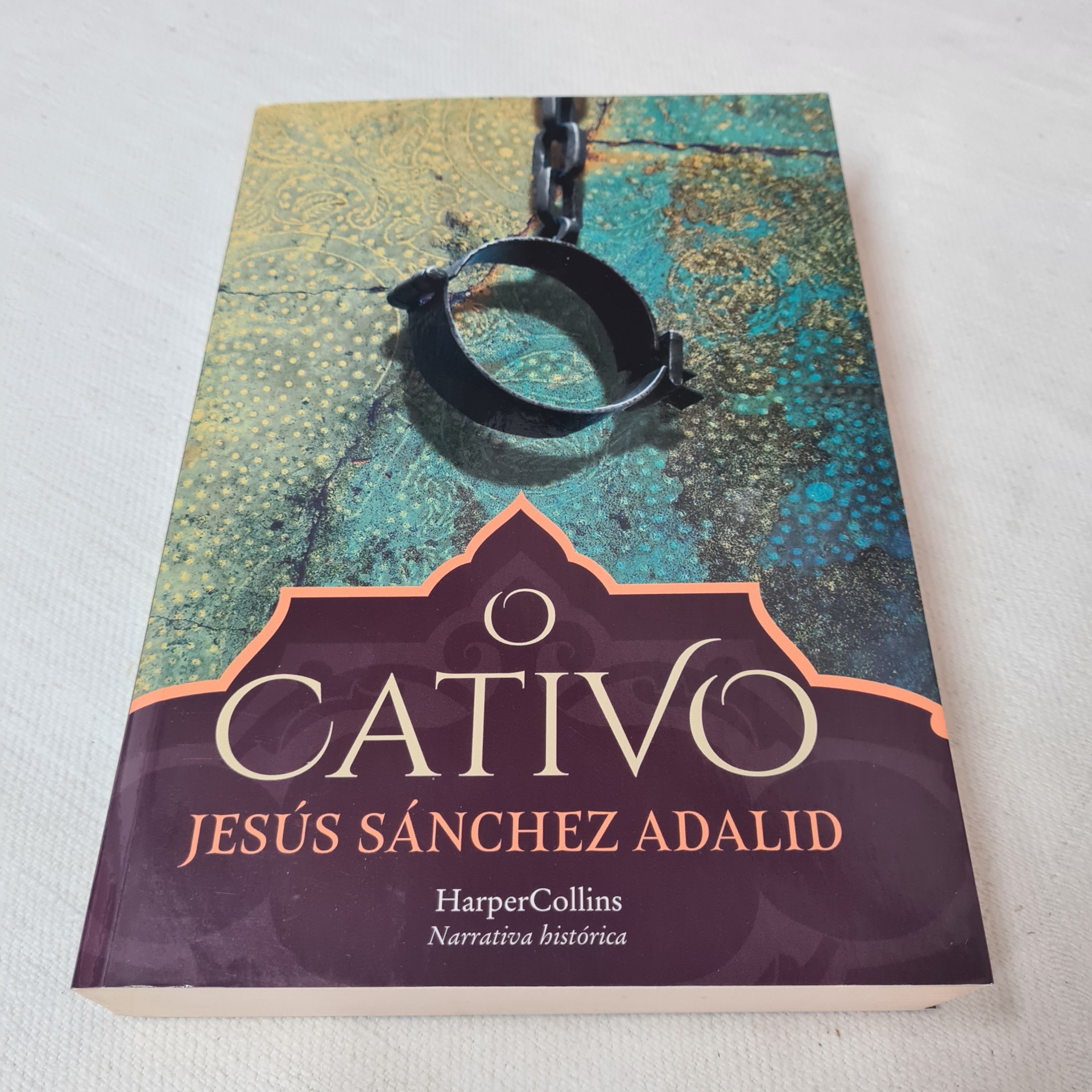 Jesús Sánchez Adalid - O Cativo Capa do livro 'O Cativo' com ilustração de manilha de ferro e corrente, fundo verde e texto em dourado