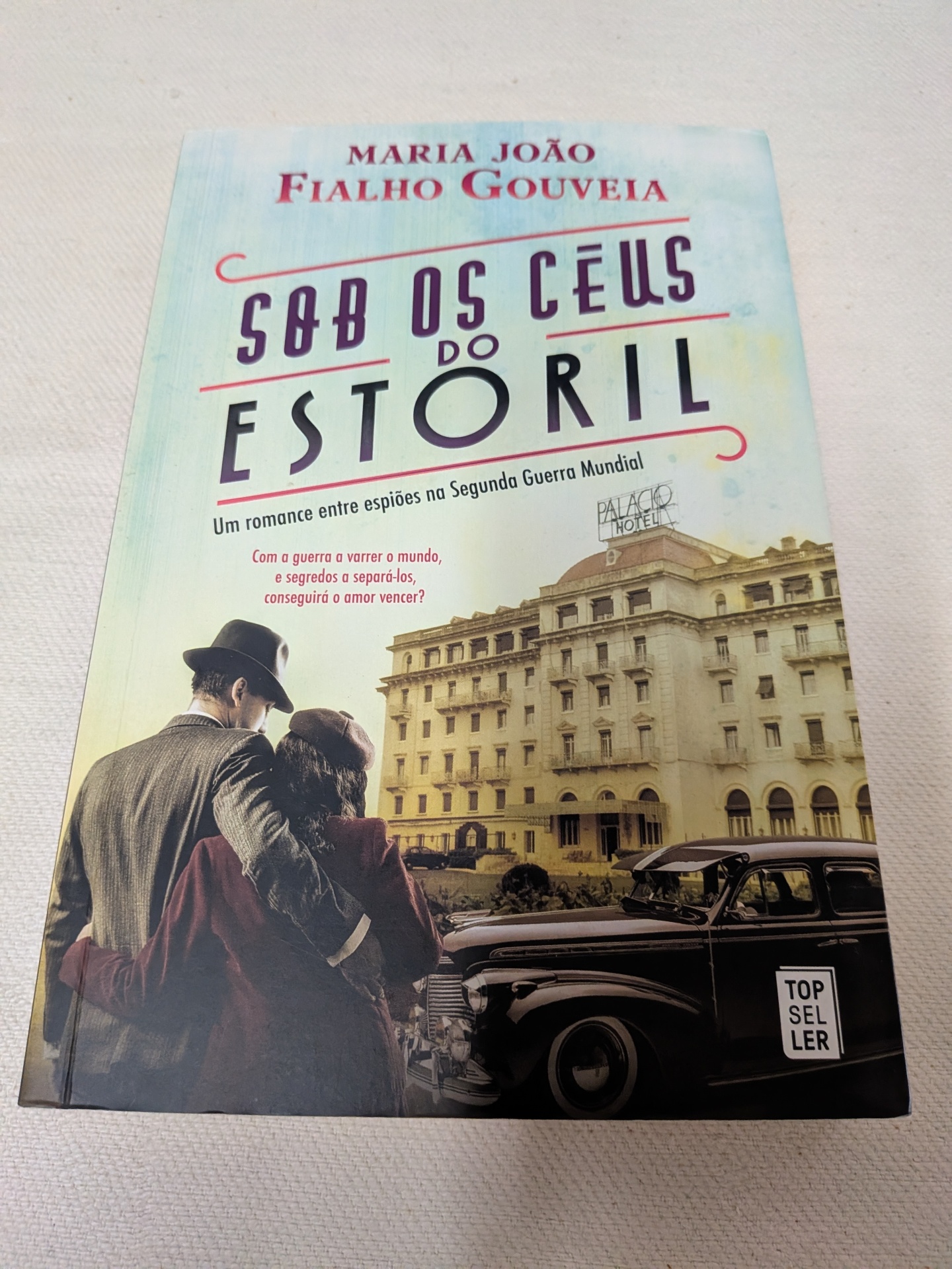 Maria João Fialho Gouveia - Sob os Céus do Estoril Capa do livro Sob os Céus do Estoril com casal vintage e carro antigo em frente a hotel.