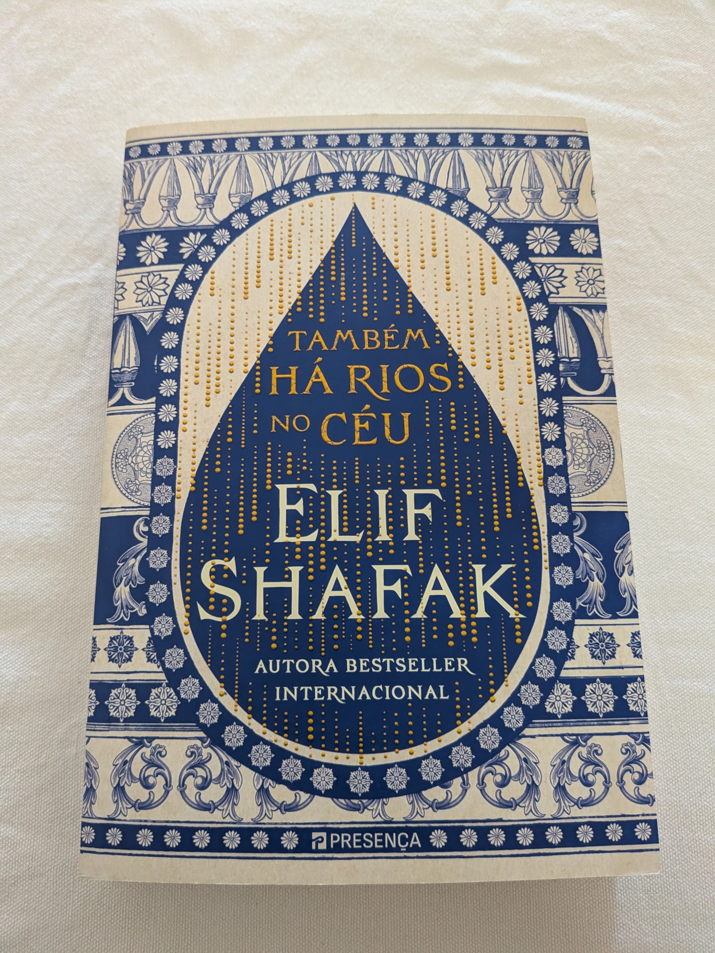 Elif Shafak - Também Há Rios no Céu Capa de livro azul e bege com detalhes florais, título e autor em relevo