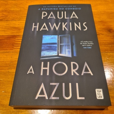 Livro 'A HORA AZUL' de Paula Hawkins com capa preta e imagem de janela para o mar