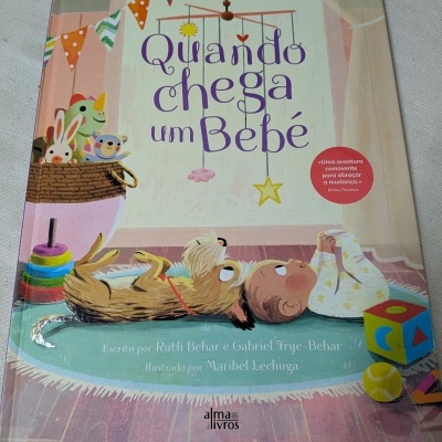 Capa de livro infantil Quando chega um Bebé com ilustração de bebé e cão deitados e brinquedos coloridos.