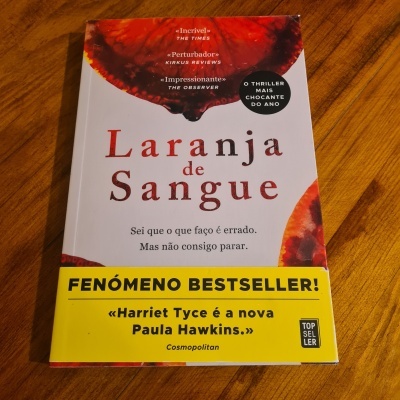 Livro 'Laranja de Sangue' com capa branca e manchas vermelhas sobre mesa de madeira