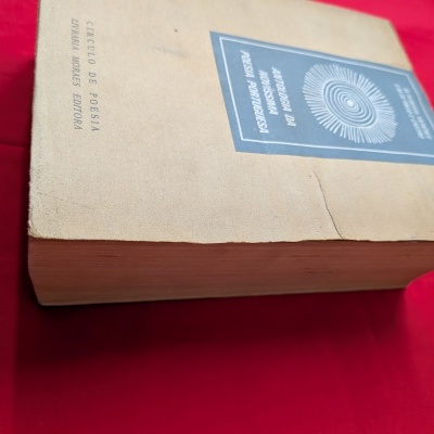 Livro antigo de capa mole castanha com etiqueta azul e texto em português sobre tecido vermelho