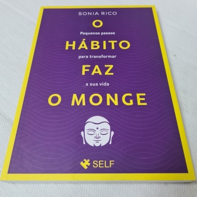 Livro 'O HÁBITO FAZ O MONGE' com capa azul e moldura amarela em superfície clara