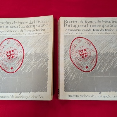 Dois livros antigos sobre História Portuguesa em capa cinzenta com carimbo vermelho, sobre tecido vermelho.