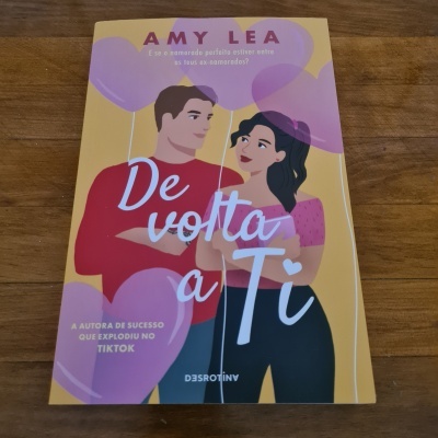 Capa do livro 'De volta a Ti' de Amy Lea com ilustração de casal e balões cor de rosa, sobre mesa de madeira.