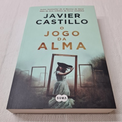 Capa do livro 'O Jogo da Alma' de Javier Castillo com mulher em vestido branco e chapéu preto em campo aberto e quadros metálicos.