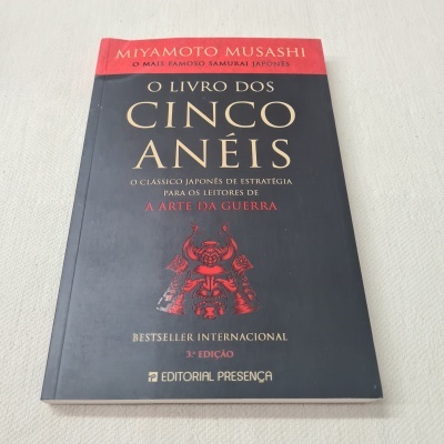 Capa do livro 'O Livro dos Cinco Anéis' de Miyamoto Musashi, com cores preta, vermelha e dourada.