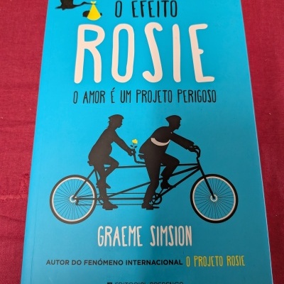 Capa azul do livro O Efeito Rosie com ilustração de uma bicicleta tandem preta com duas pessoas ao fundo vermelho