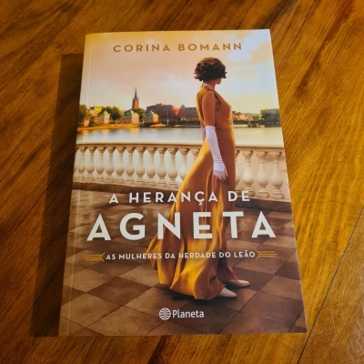 Capa do livro 'A Herança de Agneta' de Corina Bomann com imagem de mulher de vestido amarelo em varanda