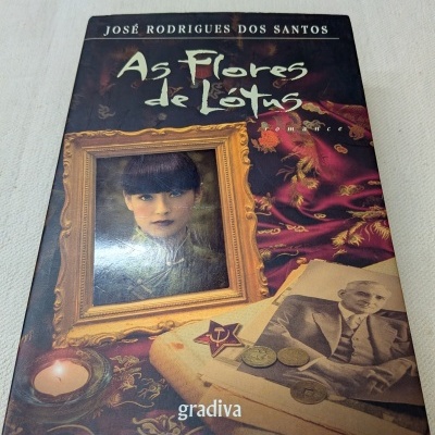 Capa do livro As Flores de Lótus de José Rodrigues dos Santos com moldura dourada, retrato de mulher e objetos antigos sobre tecido vermelho
