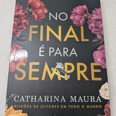 Capa de livro com título em letras amarelas e brancas, flores e anel prateado com pedra