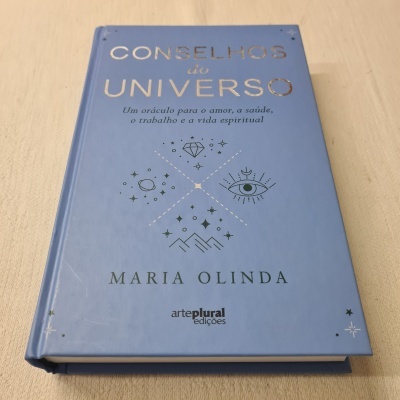 Livro azul com título 'CONSELHOS do UNIVERSO' e gráficos prateados, sobre superfície clara