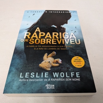 Capa do livro 'A Rapariga Que Sobreviveu' de Leslie Wolfe com imagem de uma rapariga e um ursinho de peluche