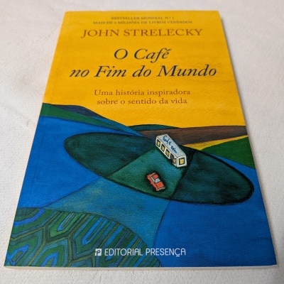 Livro 'O Café no Fim do Mundo' de John Strelecky com capa amarela e ilustração de autocarro e carro numa estrada