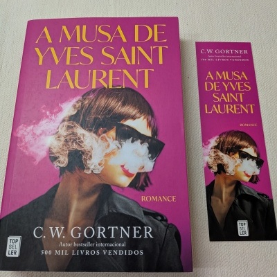 Livro 'A MUSA DE YVES SAINT LAURENT' com marcador de livro correspondente, ambos com capa rosa e imagem de mulher com óculos escuros e fumo.