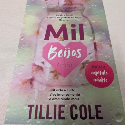 Livro Mil Beijos com capa floral e gotas de água, texto em português e selo rosa de capítulo inédito