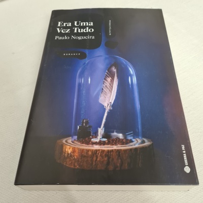 Capa de livro escura com pena branca dentro de copo de vidro sobre madeira