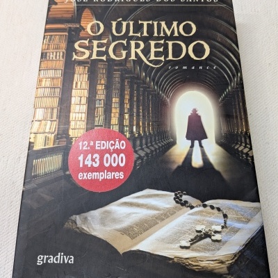 Capa do livro O Último Segredo de José Rodrigues Dos Santos com biblioteca, figura silhuetada e terço sobre livro aberto
