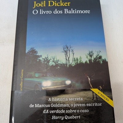 Capa do livro O livro dos Baltimore de Joël Dicker com carro antigo e veado numa estrada ao entardecer
