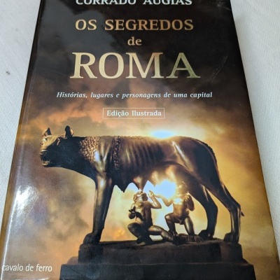 Capa do livro Os Segredos de Roma mostrando a estátua da Loba Capitolina com Rômulo e Remo em fundo de tons laranja