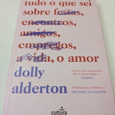 Capa de livro rosa claro com título e autor em letras vermelhas e azuis, algumas palavras riscadas a caneta azul.