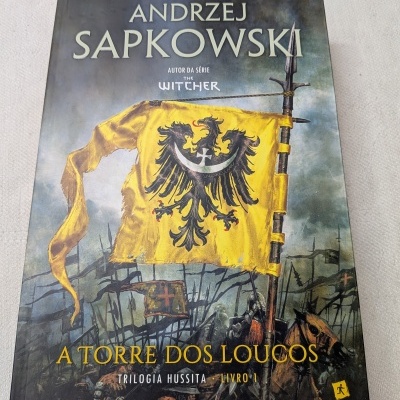 Capa do livro A Torre dos Loucos com bandeira amarela e grifo preto em cenário de batalha.
