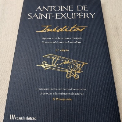 Livro em capa mole azul escuro com texto dourado e branco, incluindo o título Antoine de Saint-Exupéry Inéditos, e um desenho de avião antigo.