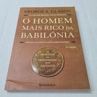 Livro O HOMEM MAIS RICO DA BABILÔNIA de George S. Clason com capa castanha e selo circular, sobre fundo branco