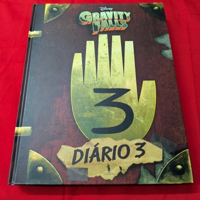 Capa do livro Disney Gravity Falls Diário 3, em tons castanho e amarelo, sobre tecido vermelho.