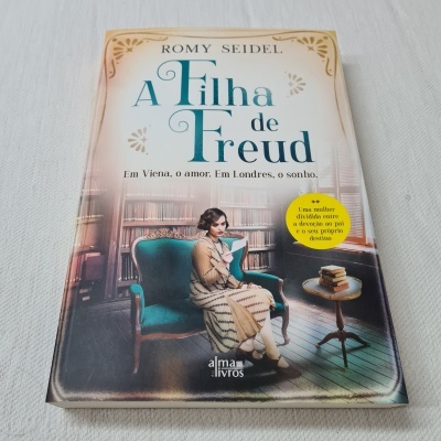 Capa do livro 'A Filha de Freud' de Romy Seidel com mulher sentada numa cadeira verde e mesa com livros.